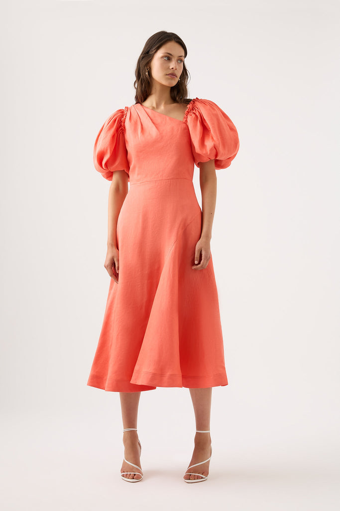 Woman wearing coral Maia Off Shoulder Midi Dress from Aje featuring relaxed skirt with curved panel seams and assymetric neckline adorned with short puff sleeves.
